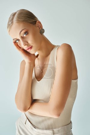 Photo for Charming blonde woman in beige tank top holding hand near perfect face and looking at camera on grey - Royalty Free Image