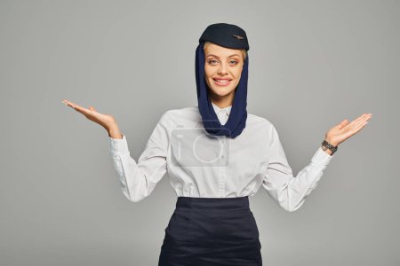 Téléchargez les photos : Joyeuse hôtesse de l'air des compagnies aériennes arabes en foulard et uniforme debout à bras ouverts sur gris - en image libre de droit
