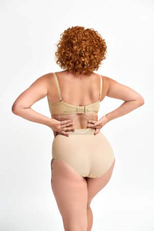plus size woman with red wavy hair posing in beige lingerie with hands on waist on white, back view
