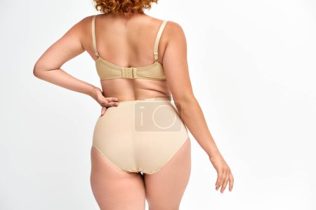 back view of curvy woman in taupe underwear posing with hand on waist on white, body positivity