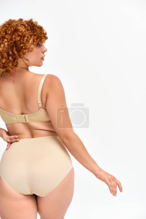 Foto de Vista trasera de pelirroja más mujer de tamaño en lencería de pie con la mano en la cadera y mirando hacia otro lado en blanco - Imagen libre de derechos