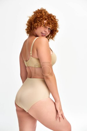 flirty woman with red wavy hair and curvy body smiling at camera in beige lingerie on white backdrop