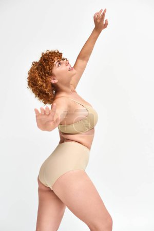 Photo for Happy plus size redhead woman in beige lingerie standing and looking up in expressive pose on white - Royalty Free Image