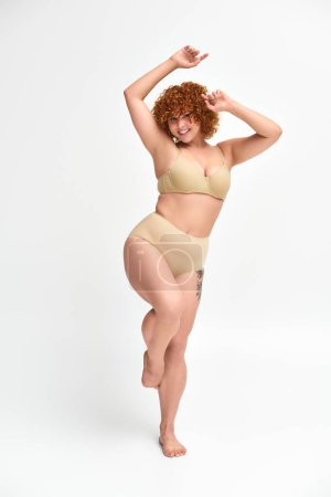 full length of jolly redhead woman with curvy body posing on one leg with raised hands on white