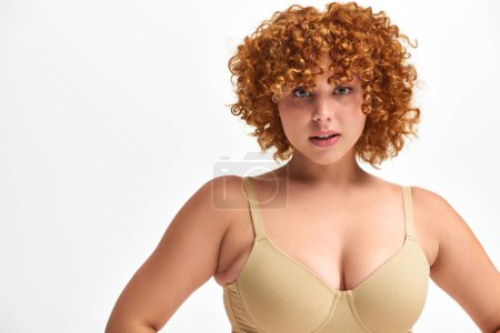 portrait of charming woman with red curly hair and curvy body and bust looking at camera on white