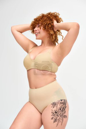 tattooed curvy woman with red curly hair posing in beige lingerie with hands behind head on white