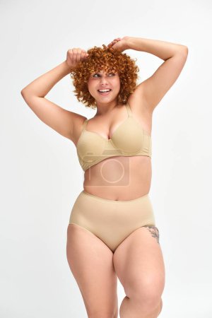 Photo for Overjoyed and redhead plus size model in beige underwear with raised hands looking away on white - Royalty Free Image