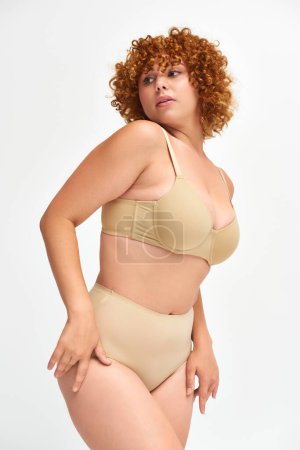 body positive redhead and curly woman in taupe underwear looking away on white, self-expression