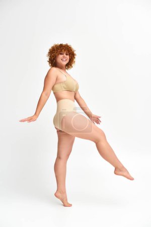 full length of happy plus size model in beige underwear standing and looking at camera on white