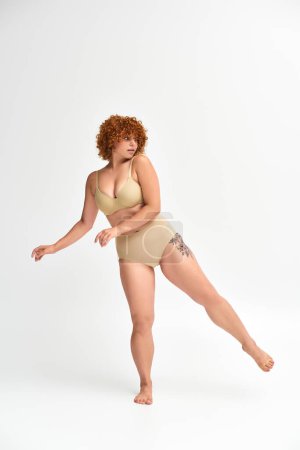 young and curvy model with tattooed hip posing in taupe lingerie on white backdrop, full length