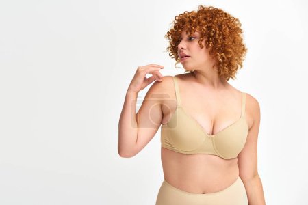 plus size woman with red curly hair in beige underwear looking away on white, non-traditional beauty