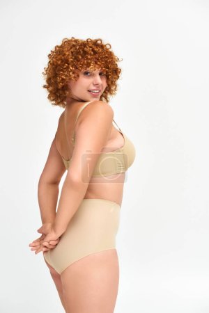 curvy and redhead woman in taupe underwear with hands behind back smiling at camera on white