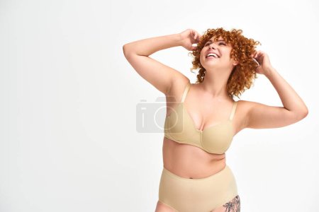 Téléchargez les photos : Joyeux et rousse plus taille femme en sous-vêtements beige souriant et regardant loin sur blanc - en image libre de droit