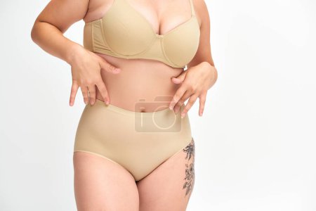 Téléchargez les photos : Vue recadrée de jeune femme plus la taille en lingerie beige avec hanche tatouée sur fond blanc - en image libre de droit