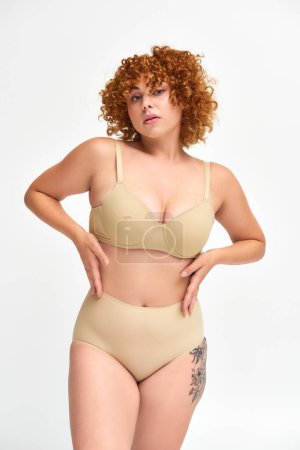 sensual redhead plus size woman in beige lingerie posing with hands on waist on white, self-esteem