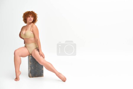 Photo for Full length of expressive redhead plus size woman in lingerie sitting on grunge wooden box on white - Royalty Free Image
