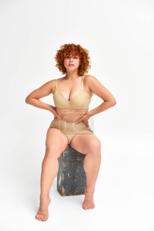 redhead curvaceous woman in beige lingerie sitting on grunge wooden box with hands on waist on white