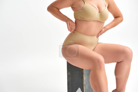 Téléchargez les photos : Vue recadrée du modèle féminin incurvé en lingerie assis sur une boîte en bois avec les mains à la taille sur blanc - en image libre de droit