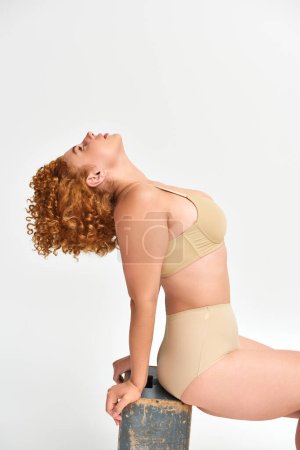 Photo for Side view of redhead plus size woman in underwear sitting and posing on wooden box on white - Royalty Free Image