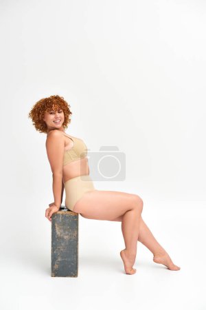 Photo for Redhead plus size woman in beige underwear sitting and smiling on wooden box on white, full length - Royalty Free Image