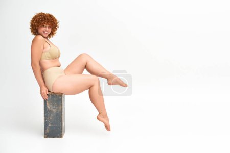 Foto de Mujer pelirroja excitada y con curvas en lencería beige sentada en caja de madera grunge en blanco, longitud completa - Imagen libre de derechos