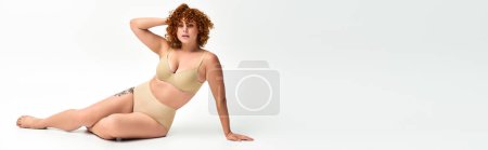 Photo for Body positive redhead woman in taupe underwear sitting on white backdrop, horizontal banner - Royalty Free Image