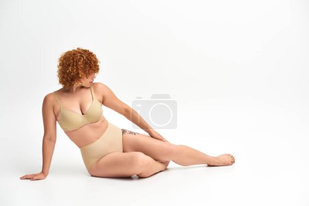 Téléchargez les photos : Pleine longueur de rousse femme avec corps courbé assis en sous-vêtements beige sur blanc, plus la beauté de la taille - en image libre de droit