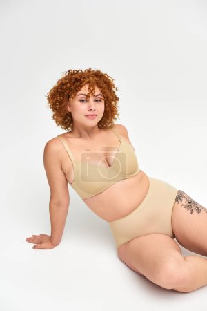 Téléchargez les photos : Femme tatouée souriante aux cheveux roux ondulés et corps taille plus assis en sous-vêtements taupe sur blanc - en image libre de droit