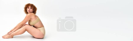 Téléchargez les photos : Femme courbée avec des cheveux roux ondulés assis dans la lingerie et regardant la caméra sur blanc, bannière horizontale - en image libre de droit