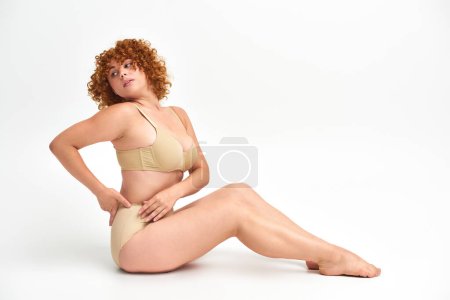 Téléchargez les photos : Pleine longueur de corps positive rousse femme en lingerie beige regardant loin tout en étant assis sur blanc - en image libre de droit