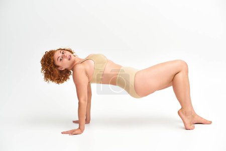 Photo for Redhead plus size woman in taupe underwear posing in reverse plank pose on white, flexibility - Royalty Free Image