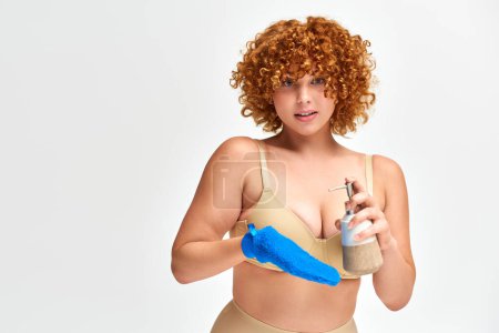 redhead plus size woman in beige lingerie with bath glove and liquid soap dispenser on white