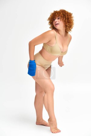 full length of redhead plus size woman washing curvy body with bath glove on white