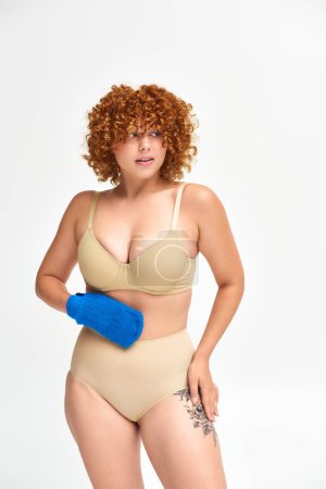 Photo for Redhead plus size woman in beige lingerie holding bath glove and looking away on white - Royalty Free Image