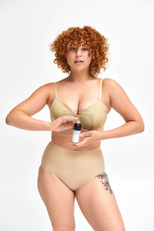 Foto de Joven mujer con curvas en lencería beige sosteniendo spray desodorante y mirando a la cámara en blanco - Imagen libre de derechos