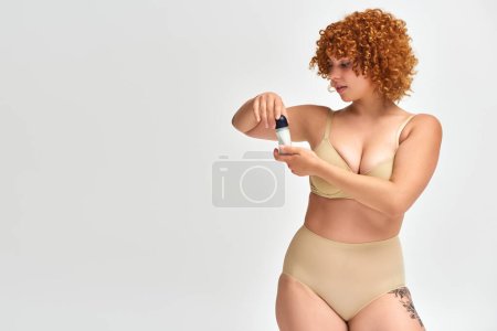 curvy woman in lingerie with red wavy hair opening roll-on antiperspirant on white, self-care ritual