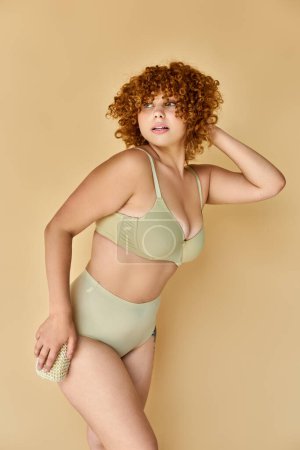 redhead plus size woman in underwear holding washcloth and looking away on white, beauty routine