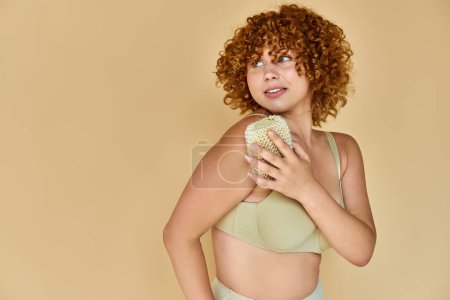 Foto de Mujer pelirroja curvilínea en ropa interior lavado cuerpo con paño y mirando hacia otro lado en beige, higiene - Imagen libre de derechos