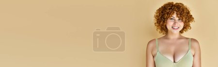 Téléchargez les photos : Jeune femme courbée aux cheveux roux ondulés posant en soutien-gorge et souriant sur fond beige, bannière horizontale - en image libre de droit