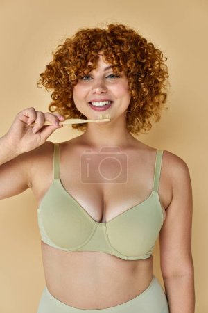 femme rousse gaie avec corps courbé tenant brosse à dents et souriant à la caméra sur fond beige