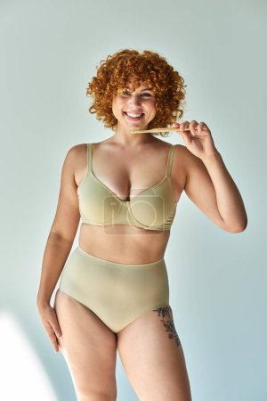 joyful redhead plus size woman in beige underwear holding toothbrush and looking at camera on beige