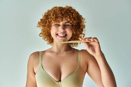 cheerful redhead and curvy woman with toothbrush smiling with closed eyes on beige backdrop