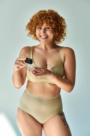 delighted and redhead plus size woman in beige lingerie holding perfume on grey background