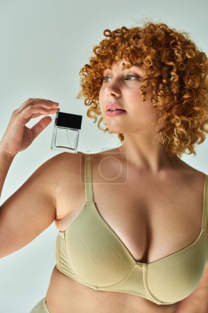 encantador modelo curvilíneo en sujetador beige con pelo rojo sosteniendo botella de perfume y mirando hacia otro lado en gris