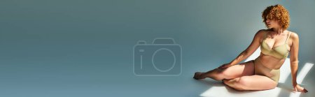 Téléchargez les photos : Sensuelle rousse plus taille femme en lingerie beige assis à la lumière du soleil sur gris, bannière horizontale - en image libre de droit