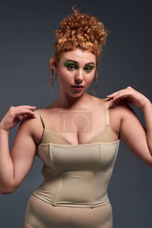 Photo for Alluring redhead woman with curvy body touching shoulder and looking at camera on dark grey - Royalty Free Image