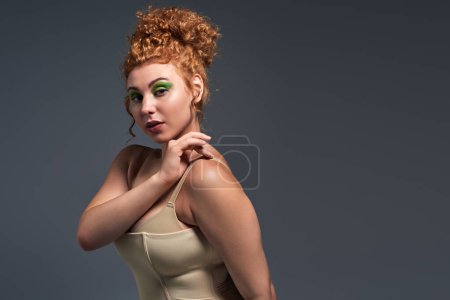 young plus size woman with bold makeup and red wavy hair touching shoulder on dark grey backdrop