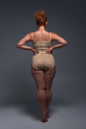 back view of curvaceous redhead woman in beige lingerie with hands on waist on dark grey backdrop