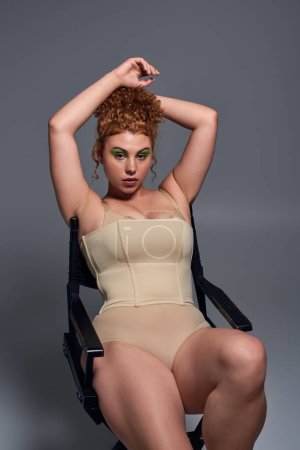 Foto de Sensual y curvilínea pelirroja en ropa interior beige sentada en silla con las manos sobre la cabeza en gris - Imagen libre de derechos