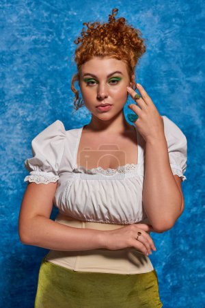 attractive and redhead plus size woman in white blouse looking at camera on blue uneven backdrop